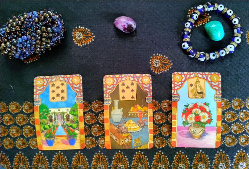 A blue linen background with gold design print. A blue and gold beaded stretch bracelet is in the upper left corner. A blue Nazar bead bracelet is in the upper right corner with a small turquoise stone in the center of it. A small purple crystal/stone is set in the middle between the two bracelets. Below the bracelets are three cards. The first depicts a garden with the number 20 and the 8 of spades from a traditional suit of cards.. The second depicts a table of food with mice on it, the number 23, and 7 of clubs from a traditional suit of cards. The third depicts a bouquet of red and white flowers, the number 9, and the Queen of spades from a traditional suit of cards.